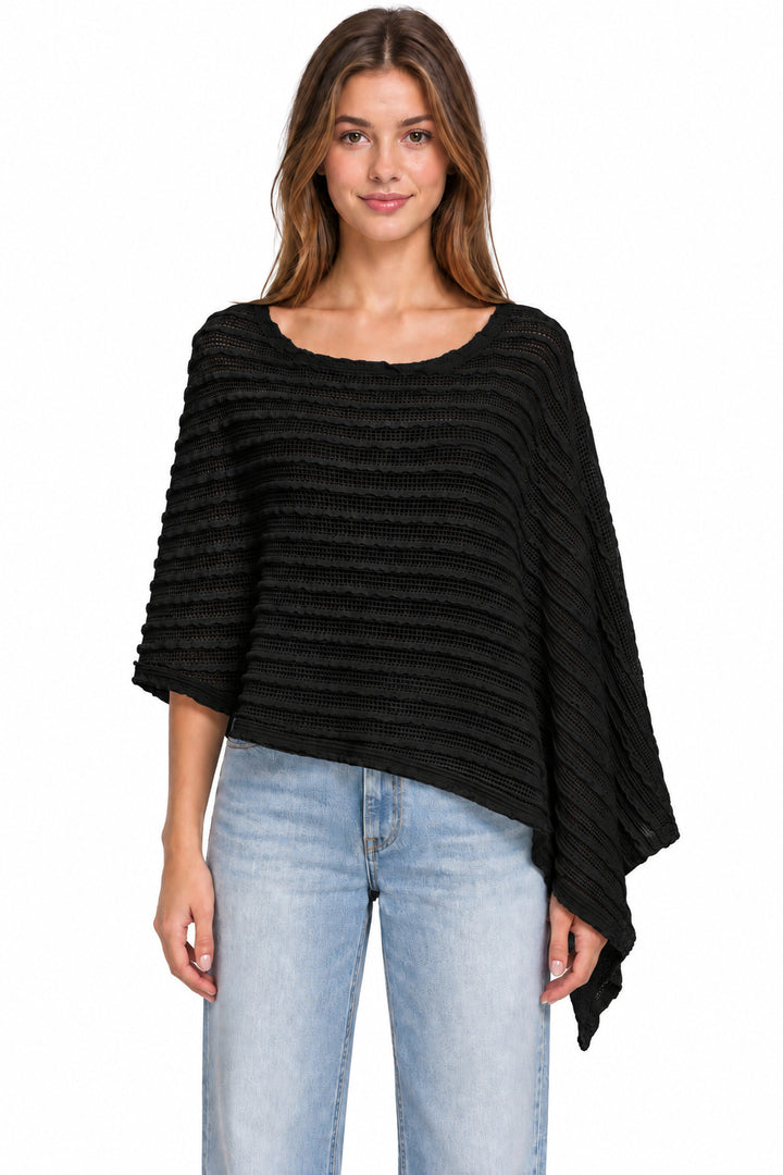 A woman modeling a black ribbed knit poncho top with a wide scoop neckline and draped sleeves, paired with light blue jeans against a plain white background.