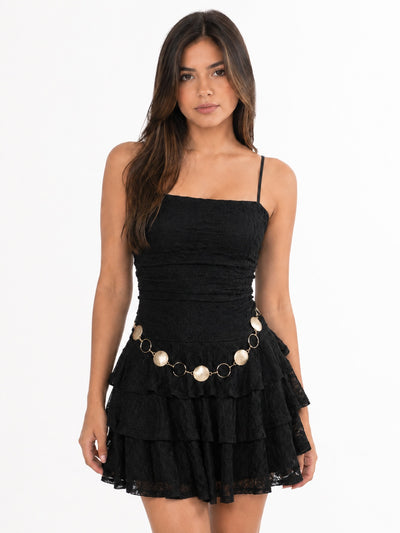 A woman modeling a black tiered mini dress with thin spaghetti straps and a decorative waist accessory featuring gold-tone rings and cream-colored oval charms, set against a plain white studio background.