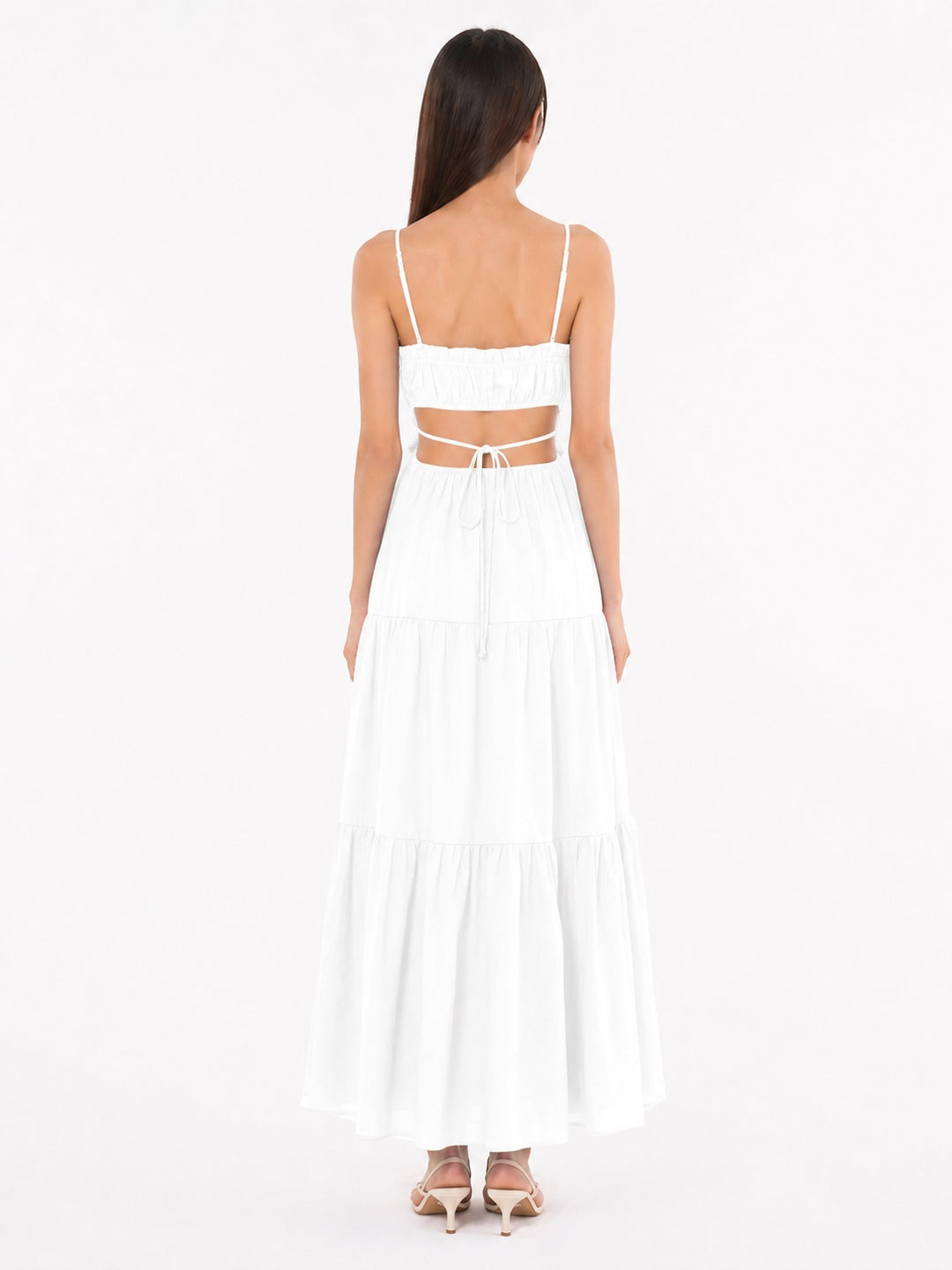 A woman viewed from the back wearing a white tiered maxi dress with thin spaghetti straps, a square neckline, and a cutout at the back tied with a string, paired with beige high-heeled sandals against a plain white studio background.