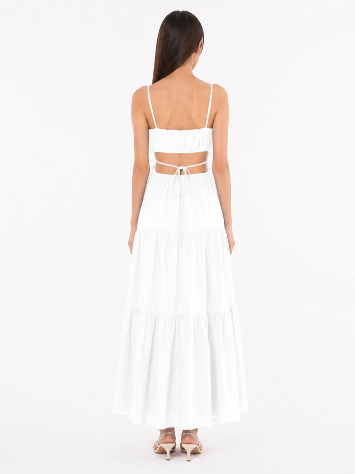 A woman viewed from the back wearing a white tiered maxi dress with thin spaghetti straps, a square neckline, and a cutout at the back tied with a string, paired with beige high-heeled sandals against a plain white studio background.