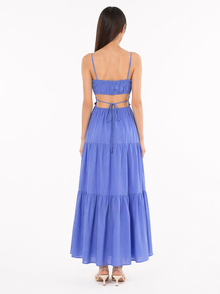 A woman viewed from the back wearing a blue tiered maxi dress with thin straps, a ruffled crop top, and a tie at the waist, paired with beige heeled sandals against a plain white background.
