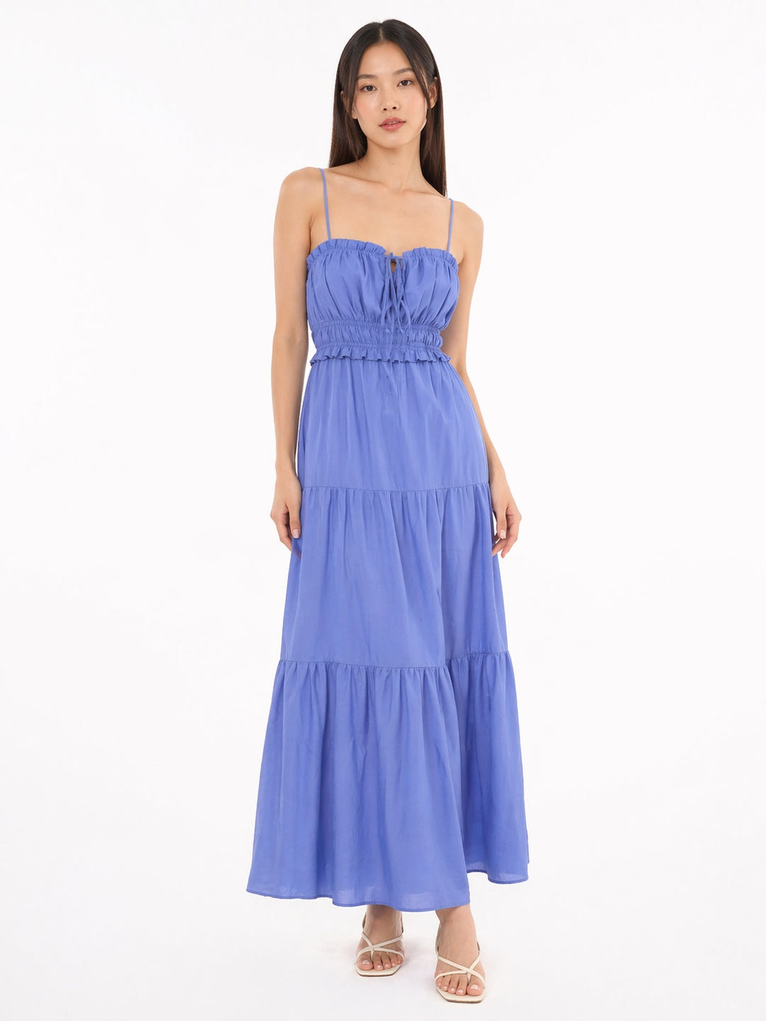 A woman modeling a light blue tiered maxi dress with spaghetti straps and a smocked bodice, standing against a plain white background.