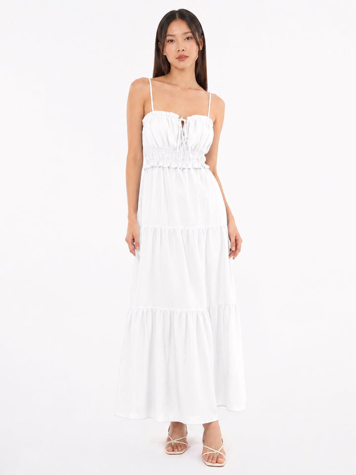 A woman modeling a white spaghetti-strap maxi dress with a smocked bodice and tiered skirt, paired with beige strappy sandals, against a plain white studio background.