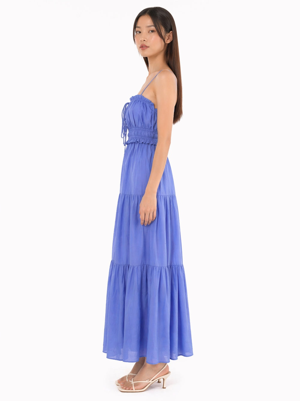 A woman modeling a strapless tiered maxi dress in a medium periwinkle blue color, shown from a side profile against a plain white studio background.