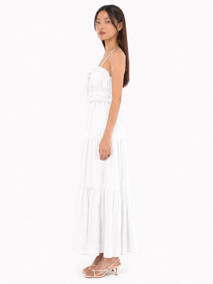 A woman modeling a white tiered maxi dress with thin spaghetti straps and a ruffled bodice, paired with beige heeled sandals, against a plain white background.